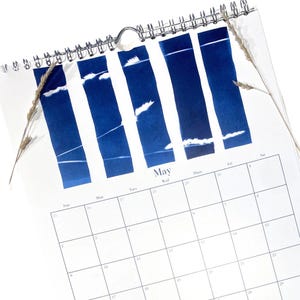 May include: A spiral-bound calendar for May, featuring a white background with blue rectangular designs and a calendar grid. The word "May" is printed in the center. Dried wheat stalks are placed on the top left and right sides.