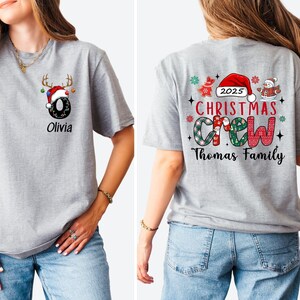 May include: A light gray Christmas-themed t-shirt. The front features a black number 0 with a Santa hat and reindeer antlers, and the name "Olivia". The back displays "2025 CHRISTMAS CREW" with festive graphics and "Thomas Family".