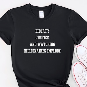 May include: Black t-shirt with white text that reads "LIBERTY JUSTICE AND WATCHING BILLIONAIRES IMPLODE." The shirt is paired with white canvas sneakers with red accents. The t-shirt is knotted at the bottom.