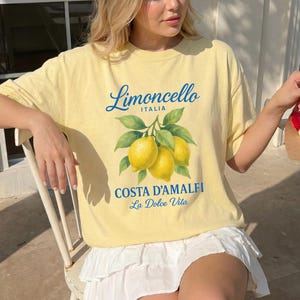 May include: A light yellow t-shirt featuring a lemon graphic with the words "Limoncello ITALIA" and "COSTA D'AMALFI La Dolce Vita" in blue. The shirt has a crew neck and short sleeves. The model is wearing a white skirt.