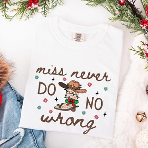 May include: White t-shirt with the text "miss never DO NO wrong" in brown script. The design features a cowboy boot and hat decorated with Christmas lights and snowflakes. The shirt is folded on a white surface.