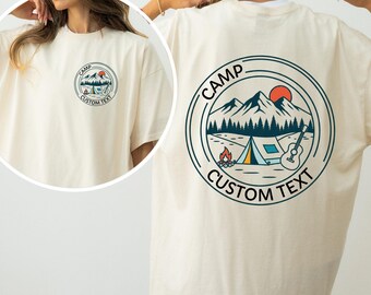 May include: Cream-coloured t-shirt with a circular graphic featuring a mountain range, tent, campfire, guitar, and the word "CAMP". The text "CUSTOM TEXT" is also included. The design is repeated on the front and back of the shirt.