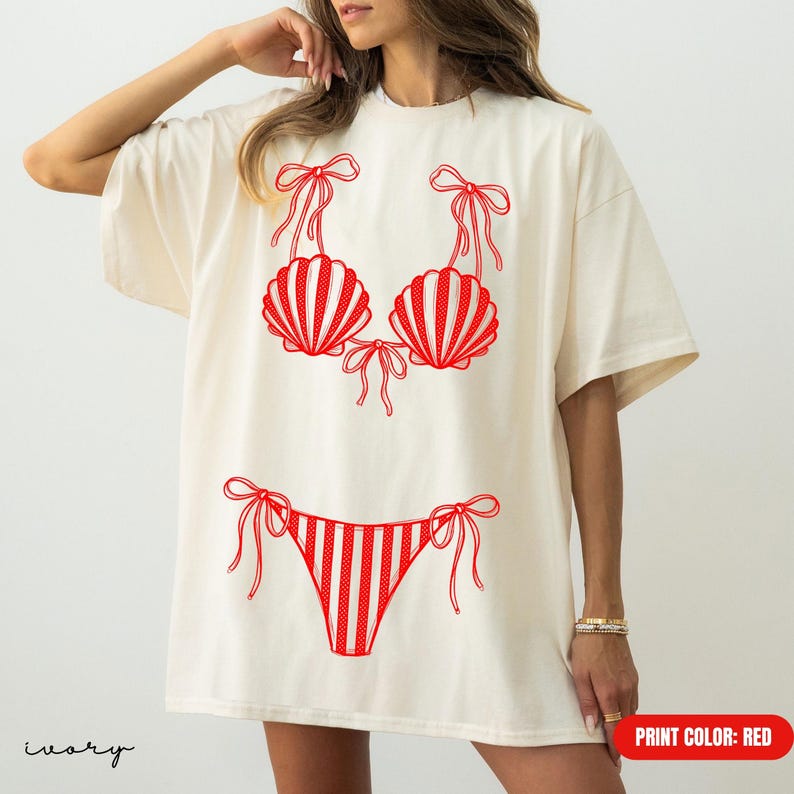 May include: An ivory short-sleeved t-shirt featuring a red graphic print of a two-piece bikini. The bikini top consists of two seashell-shaped cups with tie-string details, and the bikini bottom features a striped pattern with side ties.