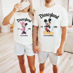 May include: Two white t-shirts with Disneyland graphics. One shirt features Minnie Mouse in pink, the other Mickey Mouse in red shorts. Both shirts have "Disneyland" in script, "est. 1955," and "California."