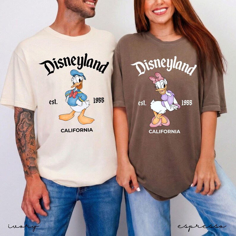 May include: Two t-shirts, one ivory and one espresso, featuring the Disneyland logo with Donald Duck and Daisy Duck. The shirts have the text "est. 1955 CALIFORNIA". The shirts are a casual style.
