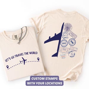Peut inclure: Deux t-shirts crème. L'un porte le texte "LET'S GO TRAVEL THE WORLD" avec un avion bleu. L'autre présente un avion bleu et des tampons de voyage. Le texte "CUSTOM STAMPS WITH YOUR LOCATIONS" est en bas.