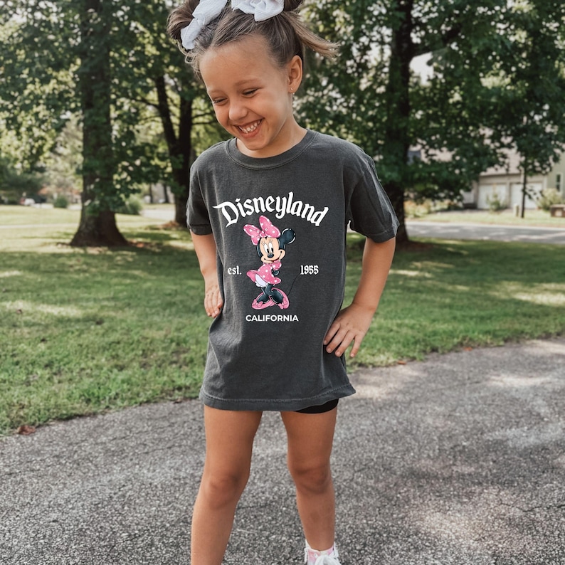 May include: Dark gray t-shirt with a Disneyland graphic featuring Minnie Mouse in a pink dress and bow. The shirt has the text "Disneyland est. 1955 CALIFORNIA" in white. The shirt is worn by a person.