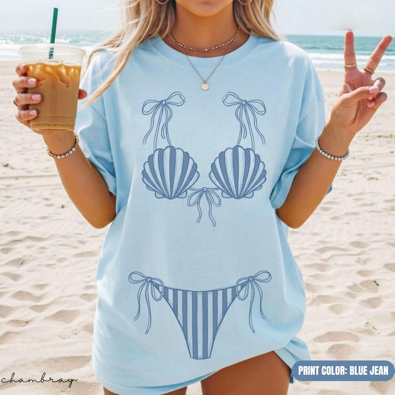 May include: A light blue t-shirt featuring a dark blue line art graphic of a bikini top made of seashells and a matching bikini bottom with tie-side details. The shirt has a crew neckline and short sleeves, showcasing a simple, casual design.