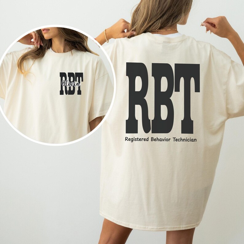 K&ouml;nnte beinhalten: Cremefarbenes T-Shirt mit den Buchstaben "RBT" in fetter schwarzer Schrift auf der R&uuml;ckseite, darunter "Registered Behavior Technician". Auf der Vorderseite steht "RBT" mit dem Namen "Olivia" dar&uuml;ber.