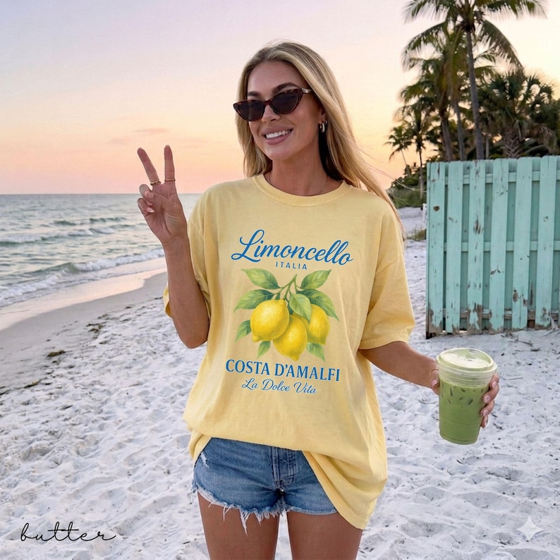 May include: Yellow t-shirt with a Limoncello Italia design featuring lemons and the text "Costa D'Amalfi La Dolce Vita". The shirt is paired with denim shorts. A person is holding a green drink on a beach.
