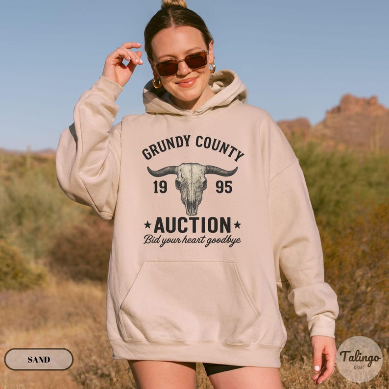 K&ouml;nnte beinhalten: Beigefarbener Kapuzenpullover mit einem Frontaufdruck &bdquo;Grundy County Auction 1995&ldquo; &uuml;ber einem Bullensch&auml;del. Der Text &bdquo;Bid your heart goodbye&ldquo; steht darunter. Der Hoodie hat eine Kapuze mit Kordelzug und eine Vordertasche.