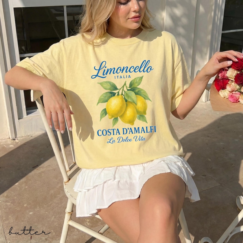 May include: A light yellow t-shirt featuring a lemon graphic with the words "Limoncello ITALIA" and "COSTA D'AMALFI La Dolce Vita" in blue. The shirt has a crew neck and short sleeves. The model is wearing a white skirt.