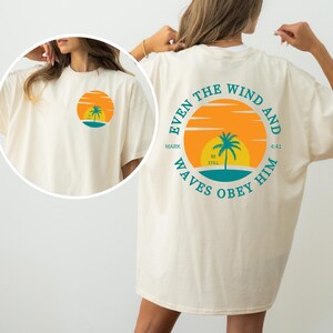 May include: A white t-shirt with a graphic on the back that says "Even the wind and waves obey him" with a palm tree and sunset graphic. The text "Mark 4:41" is also included in the graphic. The front of the shirt has a smaller version of the graphic.