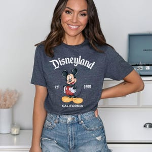 Puede incluir: Camiseta gris oscuro con el logotipo de Disneyland y un dibujo de Mickey Mouse. La camiseta tambi&eacute;n incluye el texto "est. 1955" y "CALIFORNIA".