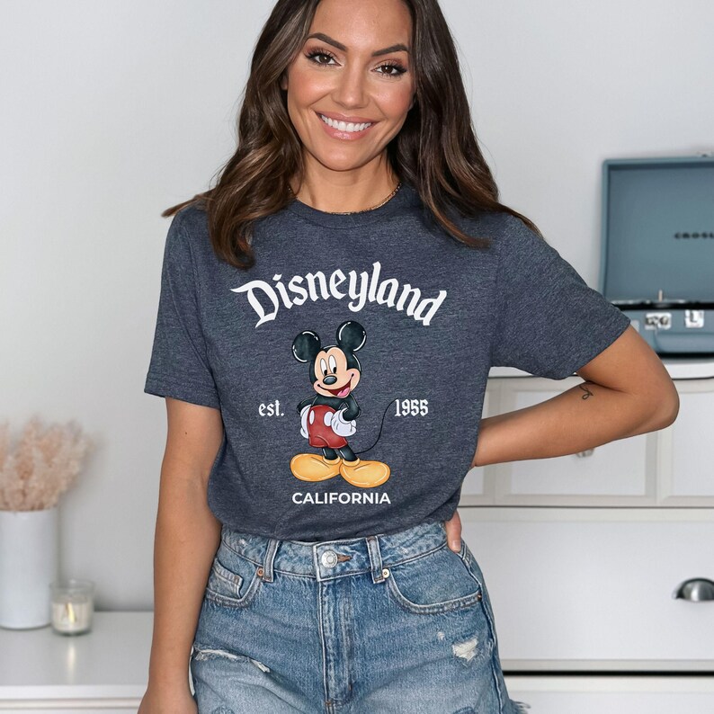 Puede incluir: Camiseta gris oscuro con el logotipo de Disneyland y un dibujo de Mickey Mouse. La camiseta tambi&eacute;n incluye el texto "est. 1955" y "CALIFORNIA".