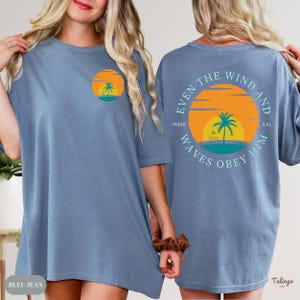May include: A light blue oversized t-shirt with a graphic of a sunset and palm tree on the front and the text "Even the wind and waves obey him" on the back. The text also includes "Mark 4:41" and "Be Still".