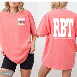 K&ouml;nnte beinhalten: Ein korallenfarbenes T-Shirt mit den Buchstaben "RBT" in Wei&szlig; auf der Vorder- und R&uuml;ckseite. Auf der Vorderseite des Shirts ist auch der Name "Emma" in einer Schreibschrift aufgedruckt. Auf der R&uuml;ckseite des Shirts stehen die Worte "Registered Behavior Technician".
