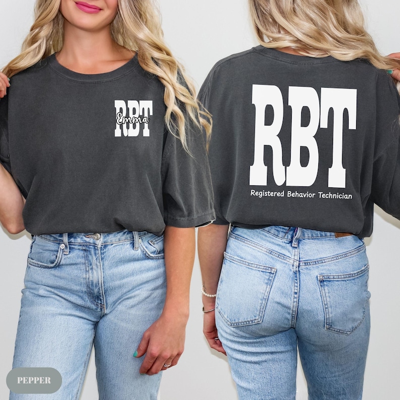 K&ouml;nnte beinhalten: Dunkelgraues T-Shirt mit den wei&szlig;en Buchstaben "RBT" auf der Vorder- und R&uuml;ckseite. Auf der Vorderseite steht der Name "Emma" in Schreibschrift unter den Buchstaben. Auf der R&uuml;ckseite steht der Text "Registered Behavior Technician" unter den Buchstaben.
