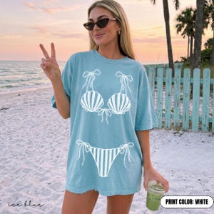 May include: An ice blue t-shirt featuring a white graphic print of a bikini set. The design includes two seashell-shaped cups with tie-string details and a matching striped bikini bottom with side ties. The shirt has short sleeves and a crew neckline.
