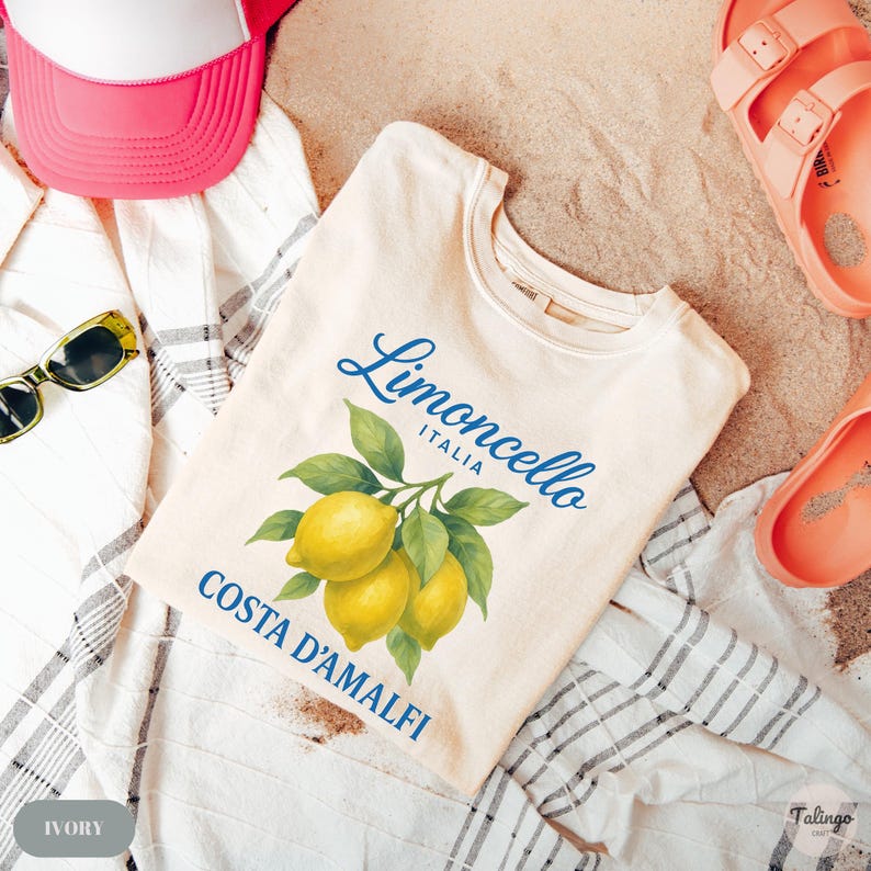 May include: Ivory-colored t-shirt with a "Limoncello Italia Costa D'Amalfi" design featuring lemons. The image includes a pink baseball cap, sunglasses, and sandals, all on a sandy surface.