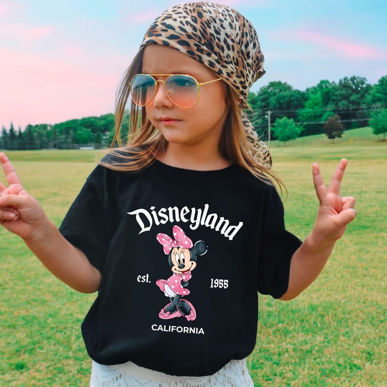 Puede incluir: Camiseta negra con el logo de Disneyland, un dibujo de Minnie Mouse y el texto "est. 1955 CALIFORNIA". La persona lleva un pa&ntilde;uelo con estampado de leopardo y gafas de sol.