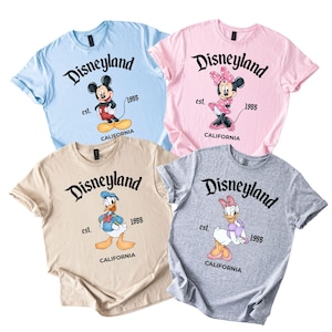 Puede incluir: Cuatro camisetas de manga corta en azul claro, rosa, beige y gris, cada una con un personaje diferente de Disney: Mickey Mouse, Minnie Mouse, Donald Duck y Daisy Duck. La palabra "Disneyland" est&aacute; impresa encima de cada personaje, con "est. 1955 CALIFORNIA" debajo.