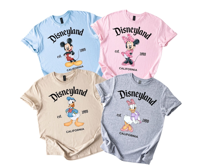 Puede incluir: Cuatro camisetas de manga corta en azul claro, rosa, beige y gris, cada una con un personaje diferente de Disney: Mickey Mouse, Minnie Mouse, Donald Duck y Daisy Duck. La palabra "Disneyland" est&aacute; impresa encima de cada personaje, con "est. 1955 CALIFORNIA" debajo.