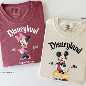 May include: Two t-shirts, one crimson and one ivory, featuring the Disneyland logo and cartoon characters. The crimson shirt displays Minnie Mouse, while the ivory shirt features Mickey Mouse. Both shirts include the text "est. 1955 CALIFORNIA".