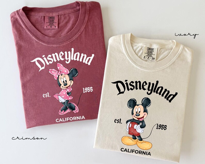 May include: Two t-shirts, one crimson and one ivory, featuring the Disneyland logo and cartoon characters. The crimson shirt displays Minnie Mouse, while the ivory shirt features Mickey Mouse. Both shirts include the text "est. 1955 CALIFORNIA".