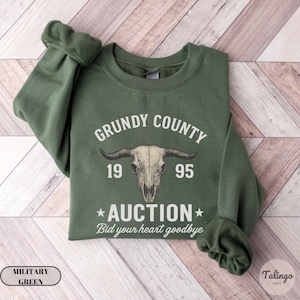 K&ouml;nnte beinhalten: Milit&auml;rgr&uuml;nes Sweatshirt mit dem Text "Grundy County Auction 1995" und einer Bullensch&auml;del-Grafik. Der Satz "Bid your heart goodbye" steht unter dem Wort "Auction". Das Sweatshirt liegt gefaltet auf einer Holzoberfl&auml;che.