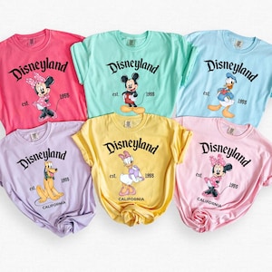 May include: A collection of six colorful t-shirts featuring various Disney characters. Each shirt displays the word "Disneyland" in a black script, along with the year "1955" and a character illustration. The shirts are in shades of pink, mint, blue, lavender, yellow, and light pink.