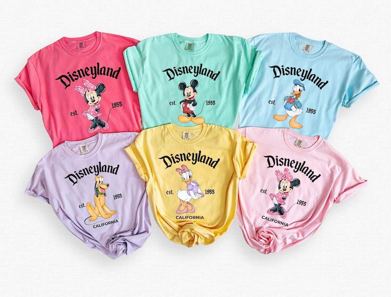 May include: A collection of six colorful t-shirts featuring various Disney characters. Each shirt displays the word "Disneyland" in a black script, along with the year "1955" and a character illustration. The shirts are in shades of pink, mint, blue, lavender, yellow, and light pink.