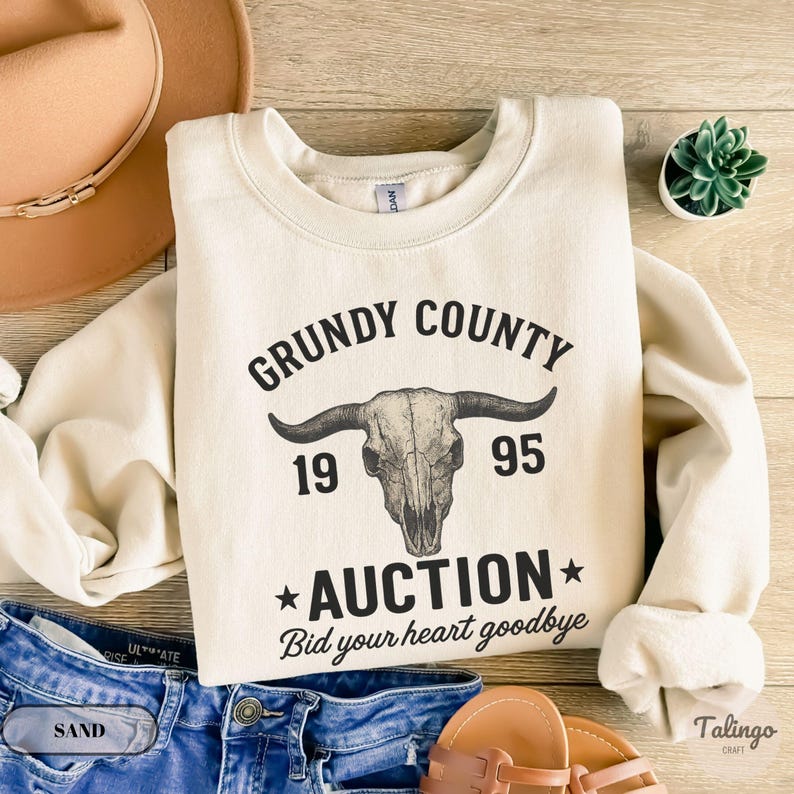 K&ouml;nnte beinhalten: Cremefarbenes Sweatshirt mit dem Text "Grundy County 1995 Auction Bid your heart goodbye" und einer Bullensch&auml;del-Grafik. Das Sweatshirt liegt auf einer Holzoberfl&auml;che mit einer Jeans, einem Hut und Sandalen.