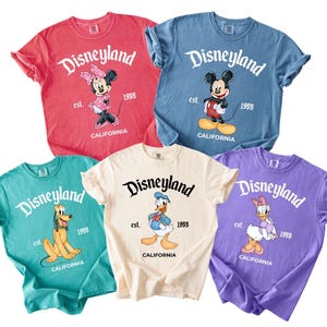May include: Five colorful t-shirts featuring Disneyland graphics. Each shirt displays a different Disney character: Minnie Mouse, Mickey Mouse, Pluto, Donald Duck, and Daisy Duck. The shirts are in red, blue, teal, cream, and purple.