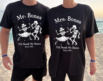 May include: Black t-shirts with white graphic of dancing skeletons, text reads "Mr. Bones" and "Mrs. Bones" above the skeletons, with "Till Death We Dance Since 2024" below. The shirts are worn on a beach.