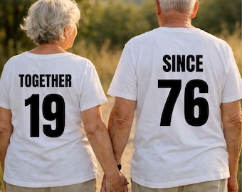 May include: A couple holding hands, wearing white t-shirts with black text. One shirt says "TOGETHER 19", the other says "SINCE 76". They are walking outdoors, with a blurred background of trees and sunlight.