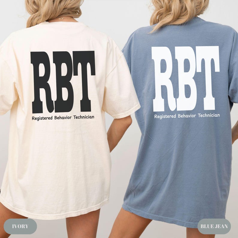 K&ouml;nnte beinhalten: Zwei T-Shirts, eines in Elfenbein und eines in Jeansblau, jeweils mit den Buchstaben "RBT" in fetter schwarzer bzw. wei&szlig;er Schrift. Unter den Buchstaben steht der Text "Registered Behavior Technician". Die Shirts sind l&auml;ssig und scheinen aus weichem Material zu sein.