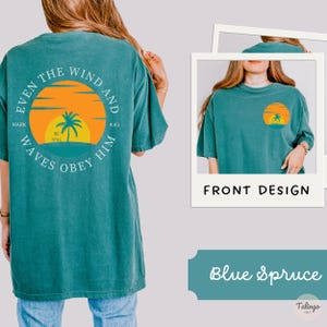 May include: A teal blue t-shirt with a graphic on the back that says "Even the wind and waves obey him" with a palm tree and sunset graphic. The front of the shirt is plain teal blue.
