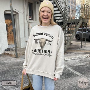 K&ouml;nnte beinhalten: Sandfarbenes Sweatshirt mit dem Text "Grundy County Auction" und einer Bullensch&auml;del-Grafik. Das Sweatshirt zeigt auch die Jahreszahlen "19" und "95" sowie den Satz "Bid your heart goodbye".