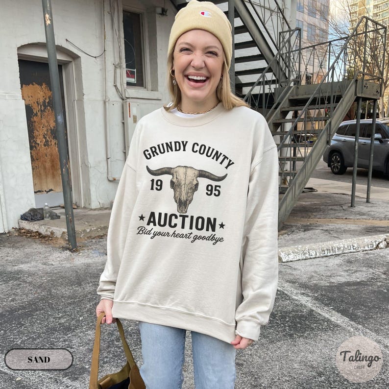 K&ouml;nnte beinhalten: Sandfarbenes Sweatshirt mit dem Text "Grundy County Auction" und einer Bullensch&auml;del-Grafik. Das Sweatshirt zeigt auch die Jahreszahlen "19" und "95" sowie den Satz "Bid your heart goodbye".