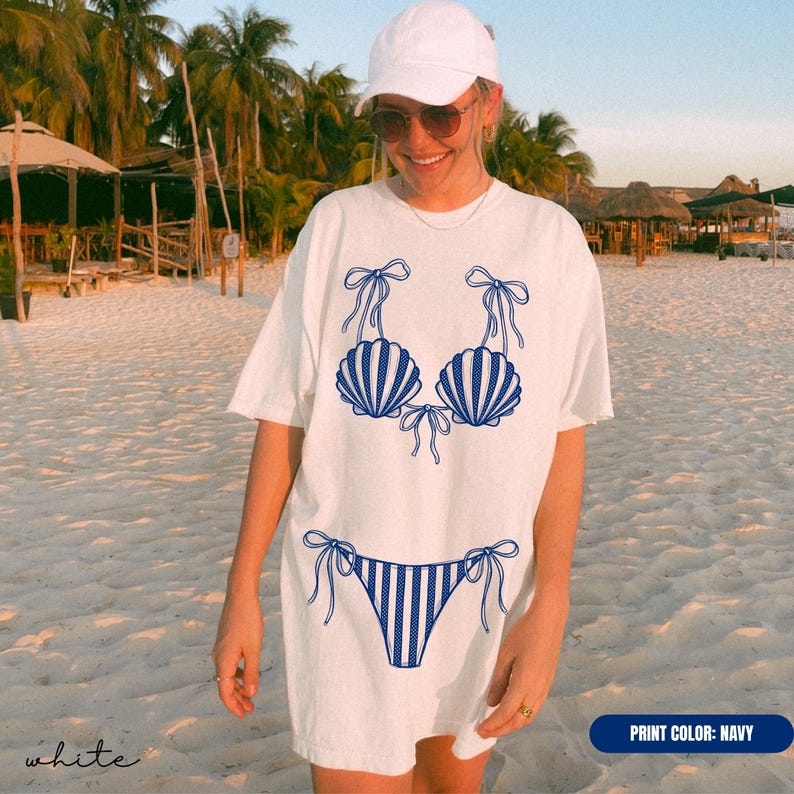 May include: A white short-sleeved t-shirt featuring a navy blue graphic print of a bikini set. The bikini top consists of two seashell-shaped cups with tie-up straps, and the bikini bottom features a striped pattern with side tie-up details.