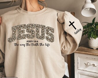 May include: Sand-colored sweatshirt with the word "JESUS" in leopard print lettering. Below the word "JESUS" is the text "JOHN 14:6" and "the way the truth the life". A black cross and the name "Ashley" are printed on the sleeve.