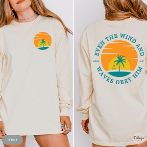 May include: Ivory long-sleeve t-shirt with a palm tree and sunset graphic on the front and the text "Even the wind and waves obey him" with a palm tree and sunset graphic on the back. The text also includes "Mark 4:41" and "Be Still".