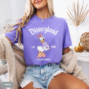 May include: Violet-colored t-shirt with the Disneyland logo and Daisy Duck graphic. The shirt features the text "Disneyland" in a white script, with "est. 1955 CALIFORNIA" below the Daisy Duck illustration. The shirt is a short-sleeved crew neck.