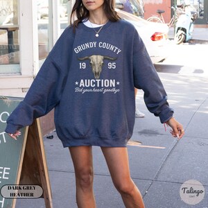 K&ouml;nnte beinhalten: Dunkelgraues meliertes Sweatshirt mit dem Text "GRUNDY COUNTY 1995 AUCTION Bid your heart goodbye" und einer Bullensch&auml;del-Grafik. Das Sweatshirt hat eine lockere Passform.