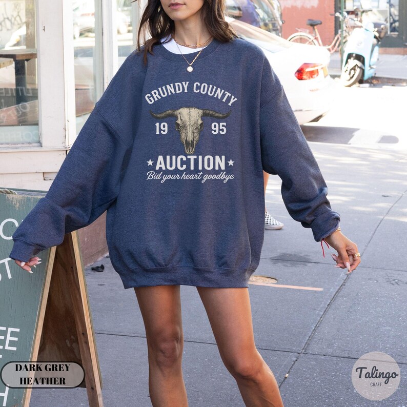K&ouml;nnte beinhalten: Dunkelgraues meliertes Sweatshirt mit dem Text "GRUNDY COUNTY 1995 AUCTION Bid your heart goodbye" und einer Bullensch&auml;del-Grafik. Das Sweatshirt hat eine lockere Passform.