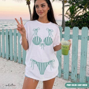 May include: A white short-sleeve t-shirt featuring a light green graphic print of a bikini set. The design includes a seashell-shaped top with tie-string details and a matching striped bikini bottom, centered on the front of the garment.