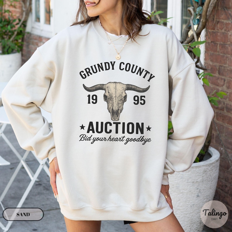 K&ouml;nnte beinhalten: Ein sandfarbener Pullover mit einem grafischen Design. Das Design zeigt den Text "Grundy County 1995 Auction Bid your heart goodbye" um einen Bullensch&auml;del. Der Pullover hat lange &Auml;rmel und einen Rundhalsausschnitt.