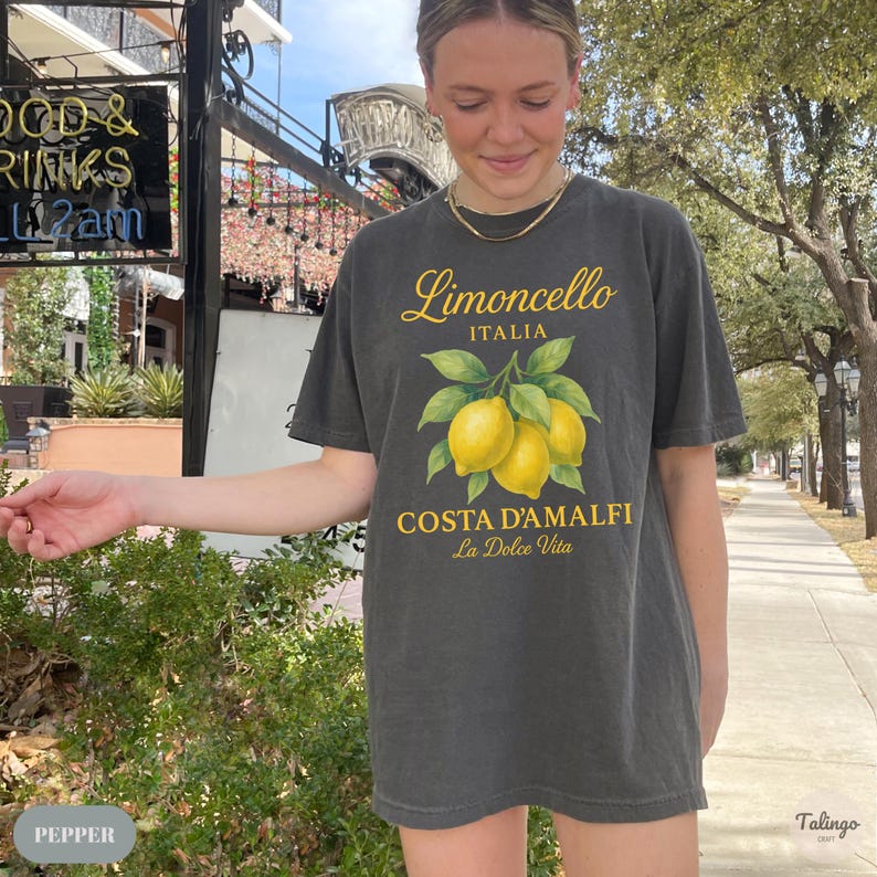 May include: Dark gray t-shirt featuring a Limoncello Italia design with yellow lemons and the text "COSTA D'AMALFI La Dolce Vita". The shirt has short sleeves and a relaxed fit. The image also shows a person wearing the shirt.