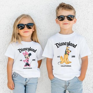 Puede incluir: Dos ni&ntilde;os con camisetas blancas con gr&aacute;ficos de Disneyland. Una camiseta presenta a Minnie Mouse, la otra a Pluto. Ambas camisetas tienen el logo de Disneyland, el a&ntilde;o 1955 y la palabra California.